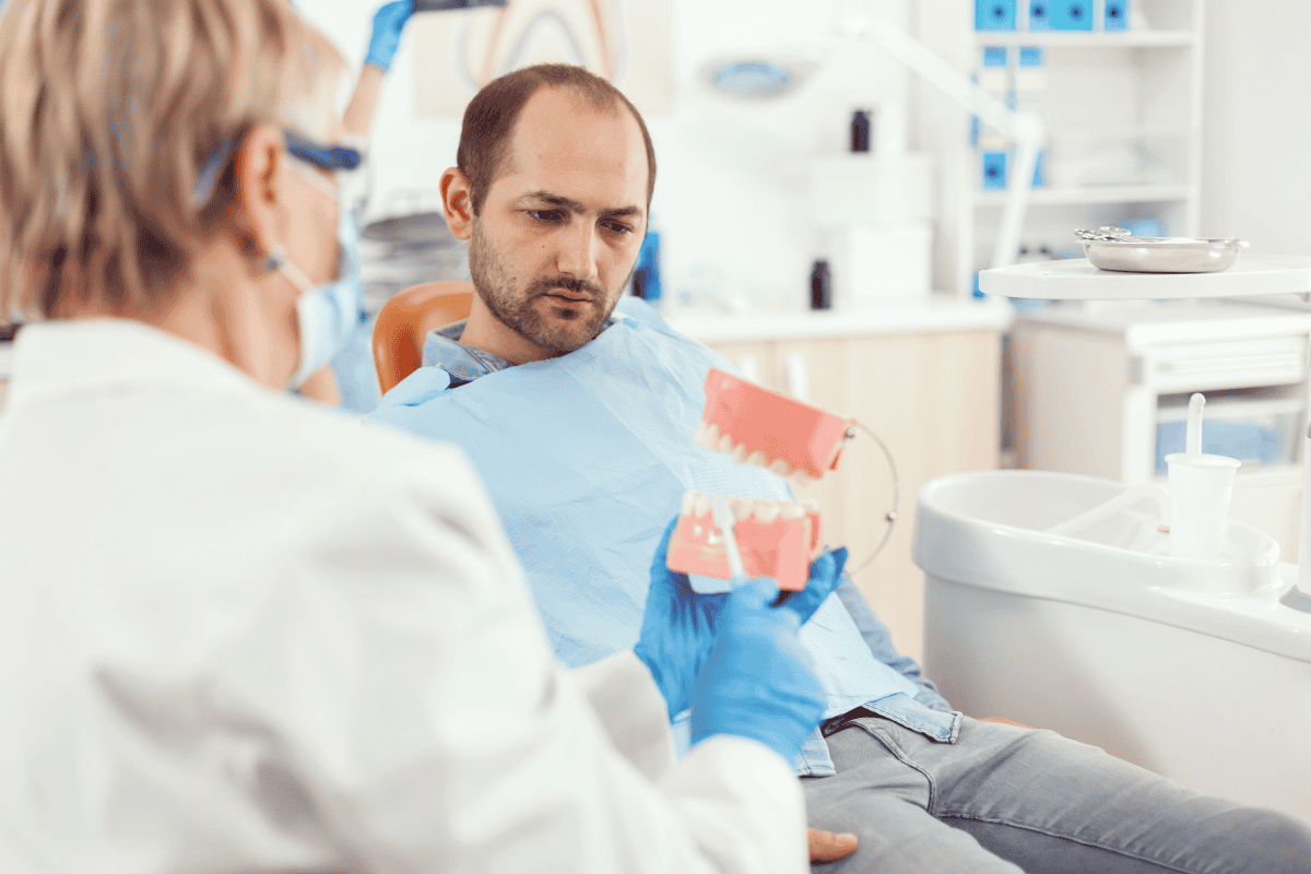 Dentist wearing gloves and a mask shows pink denture models to a patient in a bright dental clinic.