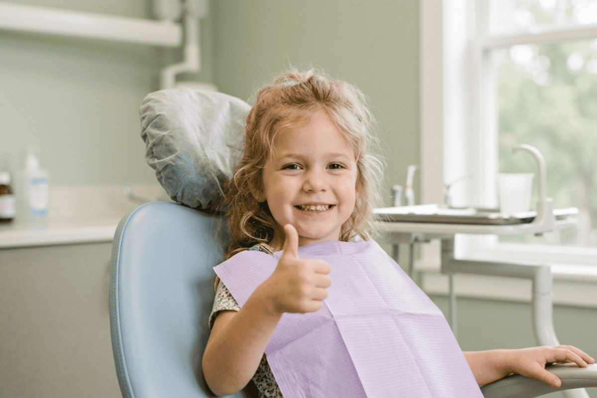 do-baby-teeth-cavities-need-to-be-filled - McDougall PhD, DDS | Tulsa Dentist Near Me | Tulsa, OK Young girl in a dental chair giving a thumbs-up, wearing a purple bib and smiling in a bright clinic setting