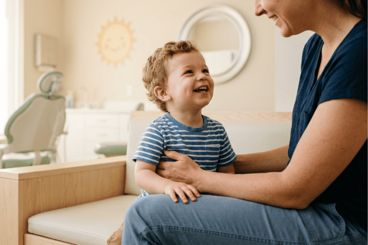 when-should-my-child-have-their-first-dentist-visit - McDougall PhD, DDS | Tulsa Dentist Near Me | Tulsa, OK Smiling toddler on a caregiver's lap in a bright pediatric dental clinic, with a dental chair in the background.